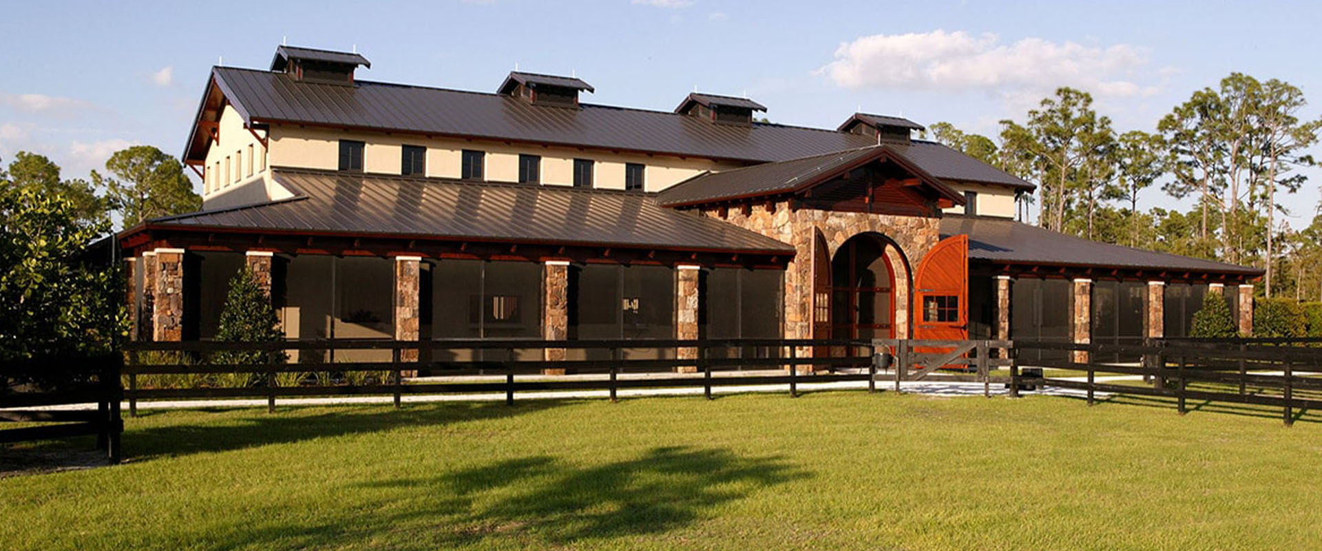 Private equestrian estate featuring a custom-designed barn with stone accents, surrounded by lush green grass and fencing, emphasizing quality and elegance in equestrian construction.